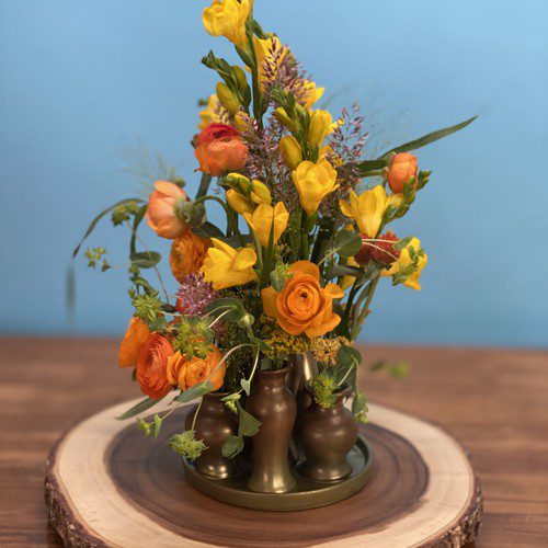 Yellow freesia and orange ranunculus in a vintage bronze bud vase on a natural wood slice, luxury florist Raleigh NC