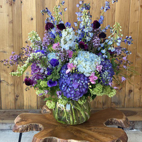 Blue and purple hydrangeas with delphinium arranged in a clear glass vase on a live-edge wood table, luxury flowers Raleigh